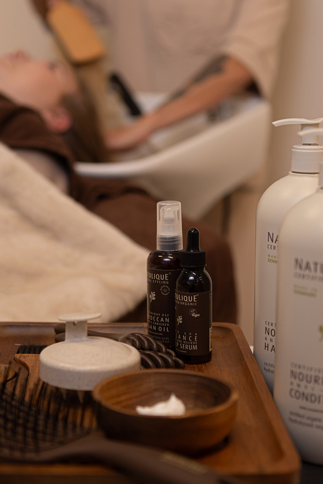 Tray met Natulique haarverzorgingsproducten tijdens een Scalp Therapy behandeling bij NSea Beauty, met op de achtergrond een ontspannen klant in een wasstoel.